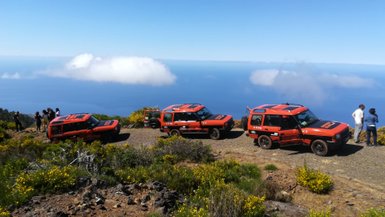 Drei orangefarbene Geländewagen stehen auf einem schmalen Weg mit Blick auf das blaue Meer und die Wolken darüber.