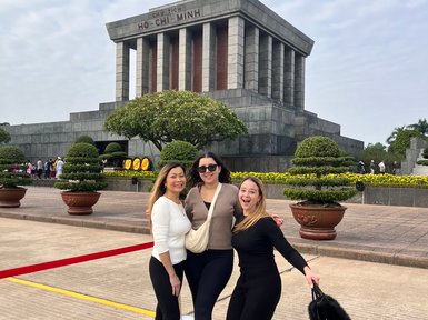 Drei Frauen posieren fröhlich vor dem Ho-Chi-Minh-Mausoleum, umgeben von gepflegten Pflanzen und einem klaren Himmel.