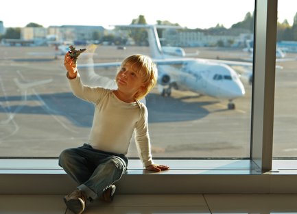 Ein kleiner Junge sitzt am Fenster eines Flughafens und hält ein Spielzeugflugzeug in der Hand, während er nach draußen schaut.