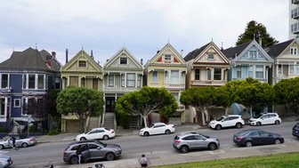 Bunte viktorianische Häuser reihen sich an einer Straße in San Francisco, umgeben von grünen Bäumen und parkenden Autos.
