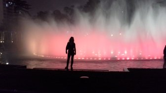 Eine silhouettierte Person steht am Rand eines beleuchteten Brunnens, während Wasserfontänen in sanften Farben sprühen.