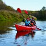 Zwei Kinder paddeln in einem roten Kajak auf einem ruhigen Fluss, umgeben von grünen Ufern und Bäumen im Hintergrund.