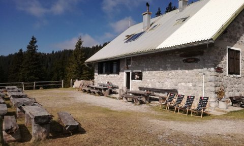 Eine gemütliche Berghütte aus Stein, umgeben von Bäumen, mit Holzstühlen und Tischen im Freien für Besucher.