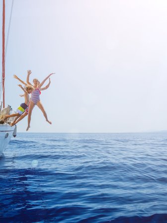 Drei Kinder springen fröhlich von einer Yacht ins glitzernde blaue Wasser, umgeben von einem klaren Himmel.