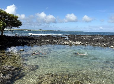 Zwei Schnorchler erkunden das klare Wasser in einer geschützten Bucht, umgeben von schwarzem Lavagestein und sanften Wellen.