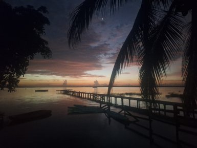 Ein ruhiger Sonnenuntergang über einem Gewässer, mit einem Holzsteg, der ins Wasser führt und Palmen im Vordergrund.