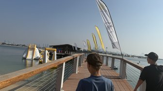 Zwei Jungen gehen auf einem Holzsteg entlang, umgeben von Wasser und bunten Fahnen, die im Wind wehen.