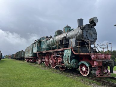 Eine alte Dampflok mit grünem und rotem Anstrich steht auf einem Gleis, umgeben von Wiesen und einem bewölkten Himmel.