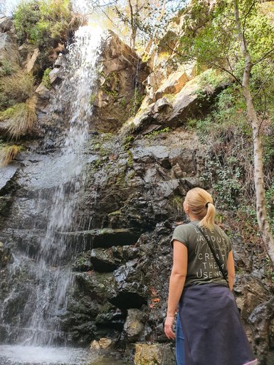 Eine Frau steht vor einem Wasserfall, der über eine felsige Klippe fließt, umgeben von üppigem Grün und Bäumen.