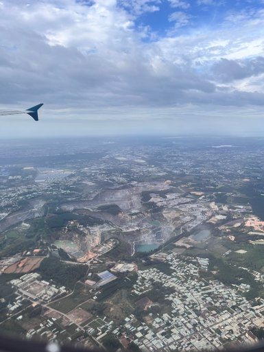 Ein Blick aus dem Flugzeug zeigt eine weite Landschaft mit grünen Feldern und städtischen Gebieten unter einem bewölkten Himmel.