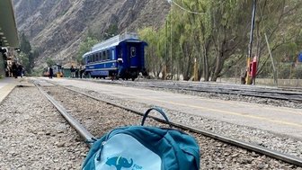 Ein blauer Rucksack steht auf den Gleisen, während ein Zug im Hintergrund an einem malerischen Bahnhof wartet.