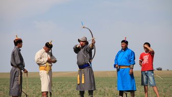 Eine Gruppe von Männern in traditioneller Kleidung, die beim Bogenschießen auf einer weiten, grünen Wiese stehen.