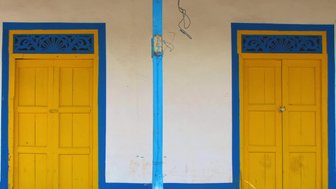 Zwei leuchtend gelbe Türen mit blauen Rahmen stehen nebeneinander an einer weißen Wand, umgeben von grünem Gras.