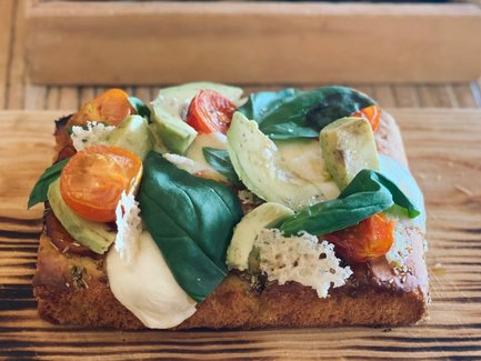 Ein frisch zubereiteter Sandwich mit Tomaten, Avocado und Spinat auf einem Holzbrett, serviert mit einer grünen Sauce.