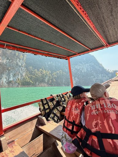 Zwei Kinder in Rettungswesten sitzen auf einem Boot und schauen auf die grüne Wasseroberfläche und die umliegenden Berge.