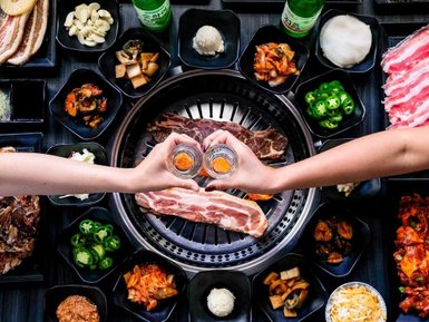 Zwei Hände halten Gläser mit einem orangefarbenen Getränk, umgeben von verschiedenen koreanischen BBQ-Gerichten auf einem Tisch.