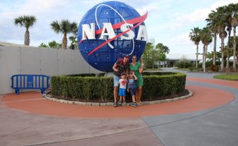 Eine Familie posiert fröhlich vor dem NASA-Symbol, umgeben von Palmen und einem gepflegten Garten.
