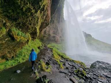 Ein Wanderer in blauer Jacke geht entlang eines feuchten Pfades, umgeben von üppigem Grün und einem beeindruckenden Wasserfall.