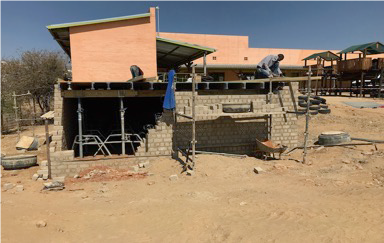 Zwei Bauarbeiter arbeiten an einer Mauer, während ein Gebäude im Hintergrund sichtbar ist, umgeben von trockenem Gelände.