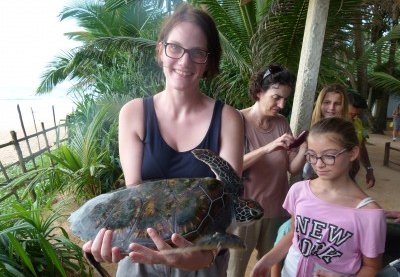 Eine Frau hält eine Schildkröte in den Händen, umgeben von grünen Pflanzen und anderen Personen im Hintergrund.