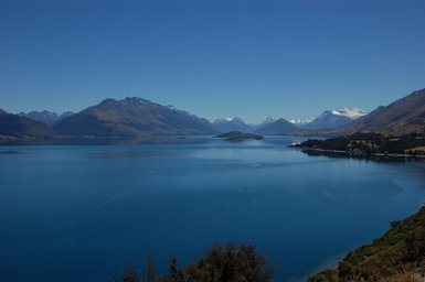 Ein atemberaubender Blick auf den klaren Lake Wakatipu, umgeben von majestätischen Bergen und strahlend blauem Himmel.