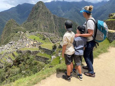 Eine Frau steht mit zwei Kindern an einem Aussichtspunkt und bewundert die Ruinen von Machu Picchu in der Ferne.