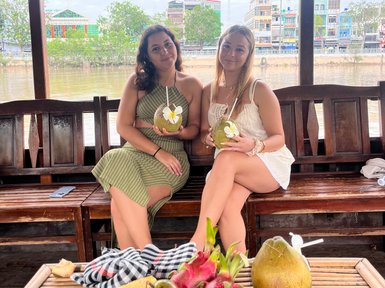 Zwei Frauen sitzen auf einem Boot im Mekong-Delta und genießen frische Früchte und Getränke, umgeben von einer malerischen Kulisse.