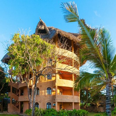 Ein mehrstöckiges Gebäude mit einem strohgedeckten Dach, umgeben von üppigem Grün und Palmen, strahlt tropisches Flair aus.