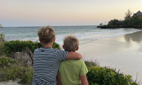 Zwei Jungen stehen am Strand, umarmt, während sie den Sonnenuntergang über dem Meer beobachten.