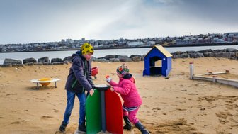 Zwei Kinder spielen auf einem Sandspielplatz, während im Hintergrund eine Stadtlandschaft und ein ruhiger Wasserbereich sichtbar sind.