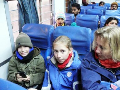 Zwei Kinder und eine Frau sitzen in einem Zugabteil, umgeben von blauen Sitzen und anderen Passagieren im Hintergrund.