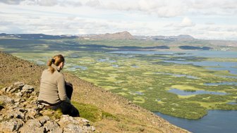 Eine Person sitzt auf einem Felsen und blickt auf die weite, grüne Landschaft mit Seen und Bergen im Hintergrund.