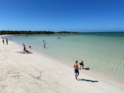 Ein sonniger Strand mit feinem, weißem Sand, wo Kinder spielen und im klaren Wasser planschen.