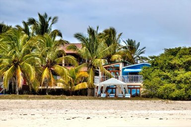 Ein tropisches Strandhaus umgeben von hohen Palmen, mit einem weißen Pavillon und blauen Liegen im Vordergrund.