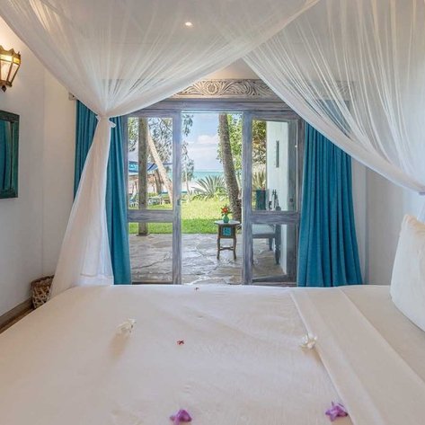 Ein helles Zimmer mit einem großen Bett, umgeben von einem Moskitonetz, bietet einen Blick auf den Strand durch große Fenster.