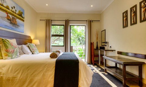 Ein gemütliches Zimmer mit einem großen Bett, bunten Kissen und Blick auf die grüne Natur durch große Fenster.