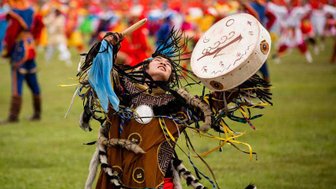 Ein junger Schamane tanzt leidenschaftlich auf einer Wiese, während er ein Trommelspiel begleitet und bunte Bänder schwingt.