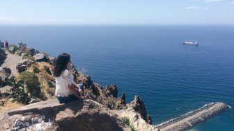 Eine Frau sitzt auf einem Felsen und blickt auf das ruhige blaue Meer, während ein Schiff in der Ferne vorbeifährt.