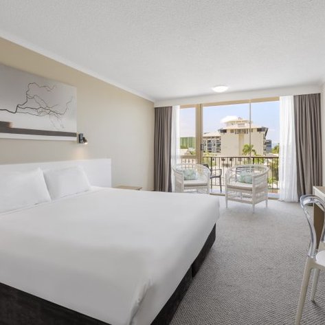 Ein modernes Hotelzimmer mit einem großen Doppelbett, hellem Teppich und einer Fensterfront, die auf einen Balkon führt.