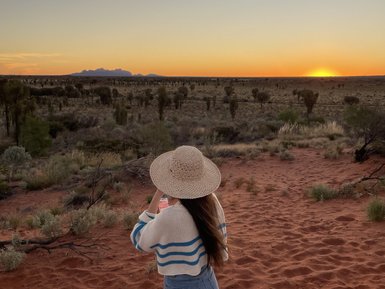 Eine Person mit einem Strohhut steht auf rotem Sand und genießt den Sonnenuntergang über der weiten, trockenen Landschaft.