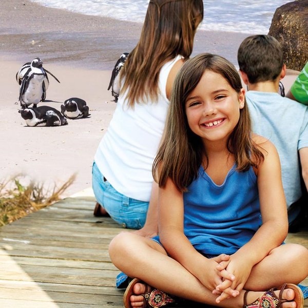Ein fröhliches Mädchen sitzt auf einem Holzsteg und lächelt, während ihre Familie und Pinguine im Hintergrund zu sehen sind.