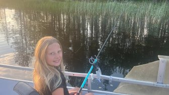 Ein Mädchen sitzt auf einem Boot am See, hält eine Angelrute und lächelt, umgeben von Bäumen und ruhigem Wasser.