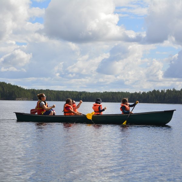 Eine Gruppe von vier Personen paddelt in einem Kanu auf einem ruhigen See, umgeben von üppigem Wald und bewölktem Himmel.