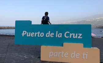 Ein Junge sitzt auf einem bunten Schild mit der Aufschrift 'Puerto de la Cruz' am Strand, während das Meer im Hintergrund glitzert.
