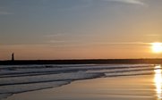 Ein malerischer Sonnenuntergang über dem Meer, mit sanften Wellen und einem Leuchtturm in der Ferne.
