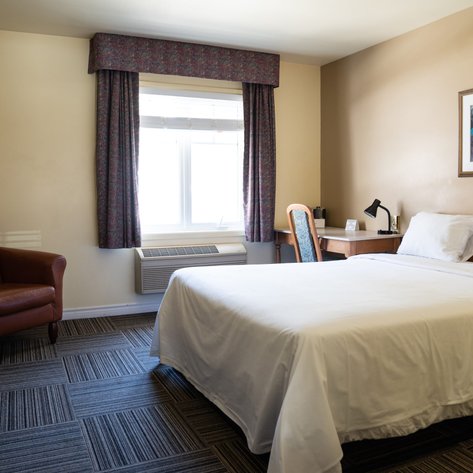 Ein einfaches Hotelzimmer mit einem großen Bett, einem Schreibtisch und einem gemütlichen Sessel in warmen Farbtönen.