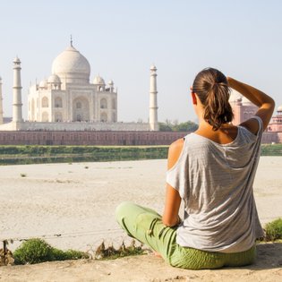 Eine Frau sitzt am Ufer und betrachtet das majestätische Taj Mahal, umgeben von einer ruhigen Landschaft.
