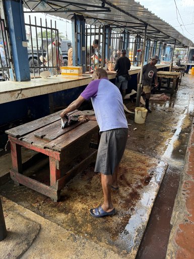 Ein Mann schneidet Fisch an einem Marktstand, während andere Verkäufer im Hintergrund beschäftigt sind.