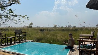 Ein Junge sitzt am Rand eines Pools und spielt mit Wasser, umgeben von hohen Gräsern und einem klaren Himmel.