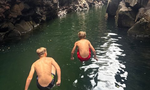 Zwei Jungen springen gleichzeitig in ein grünes Gewässer, umgeben von steilen, felsigen Wänden und üppigem Grün.